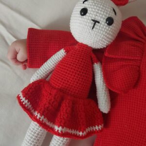 Adorable crochet rabbit doll in red outfit next to a baby's hand on a red blanket.