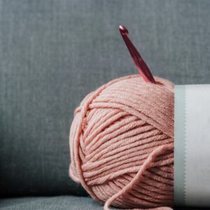 Ball of soft yarn for knitting with pink crochet hook on gray comfortable couch at home